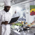 Dedicated african american chef does research on laptop to cook gourmet dishes, working in restaurant kitchen to create delicious fine dining meals. Young man doing food prep.